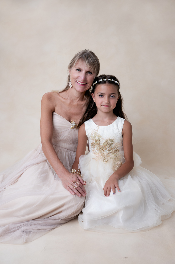 Mother and 9 year old daughter posing indoors on cream color and white dresses, respectively.