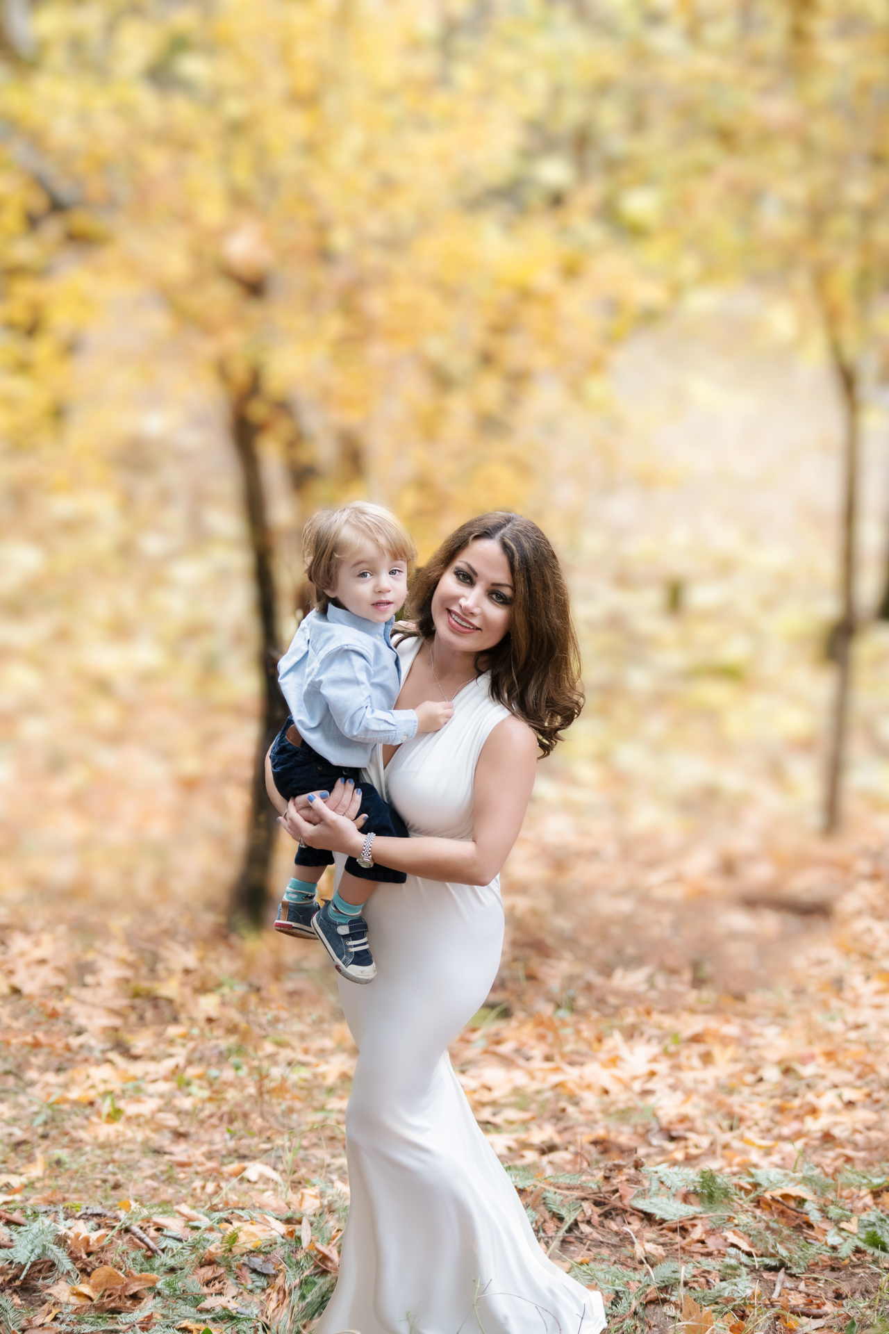 Mom wearing a white dress holding her 2 year old son posing outdoors during fall season.