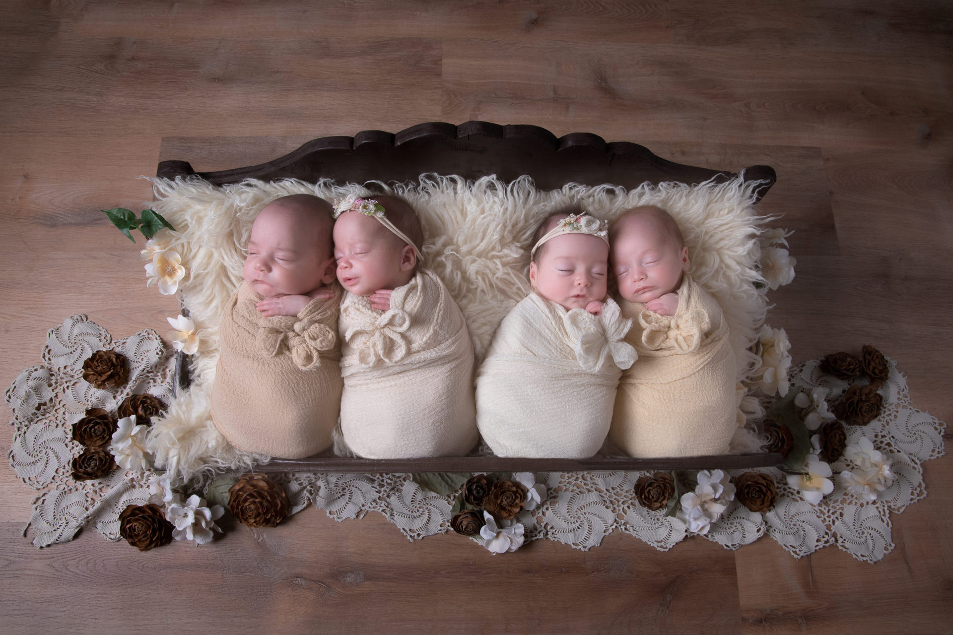 4 newborns siblings rest on a wood bed prop. 2 of them wearing light colored headbands and wraps. Two of them on darker brown wrap tones. pineapples decorate the scene. 90 degrees angle shot.
