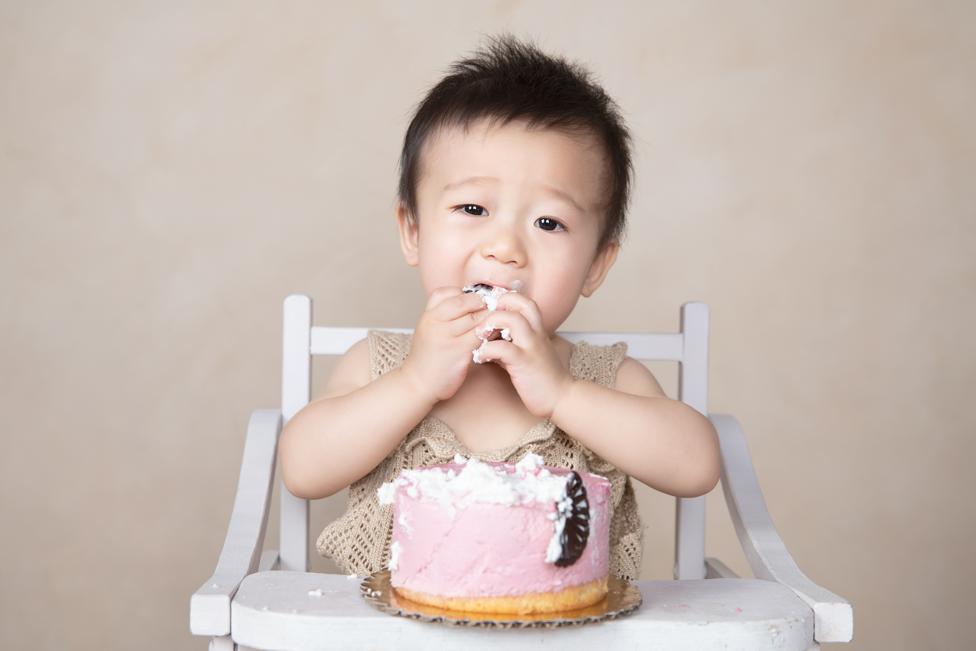 Baby boy sits on chair while eating a piece of cake that sits in front on him in the attached chair table. Light brown backdrop