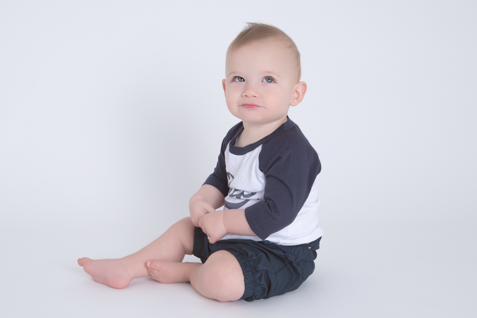 Baby boy wearing blue outfit sitting down looks up. White backdrop