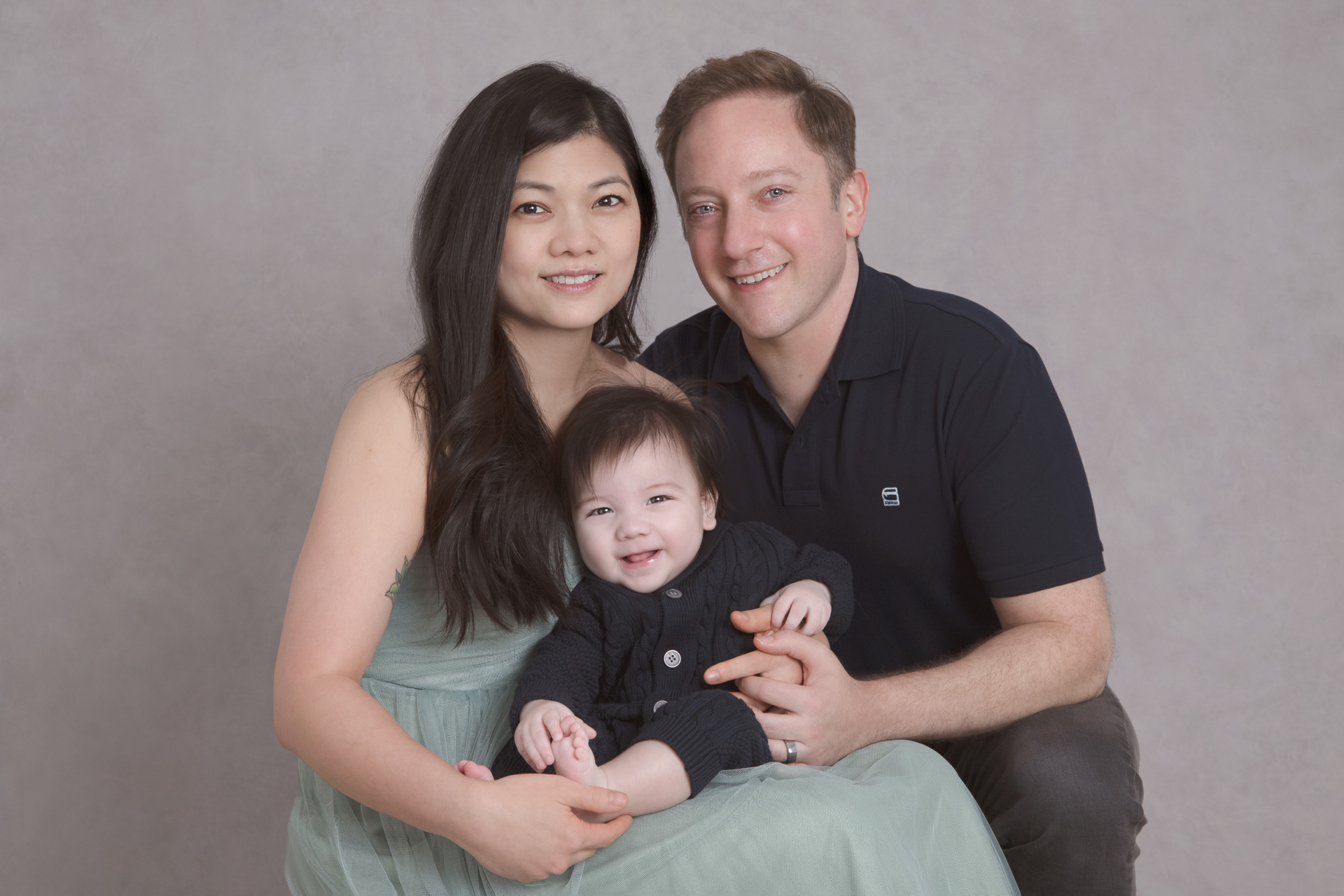 Couple looking at the camera while holding their baby boy son. Grey backdrop. Boys on dark blue tones, mom wears a light green dress.