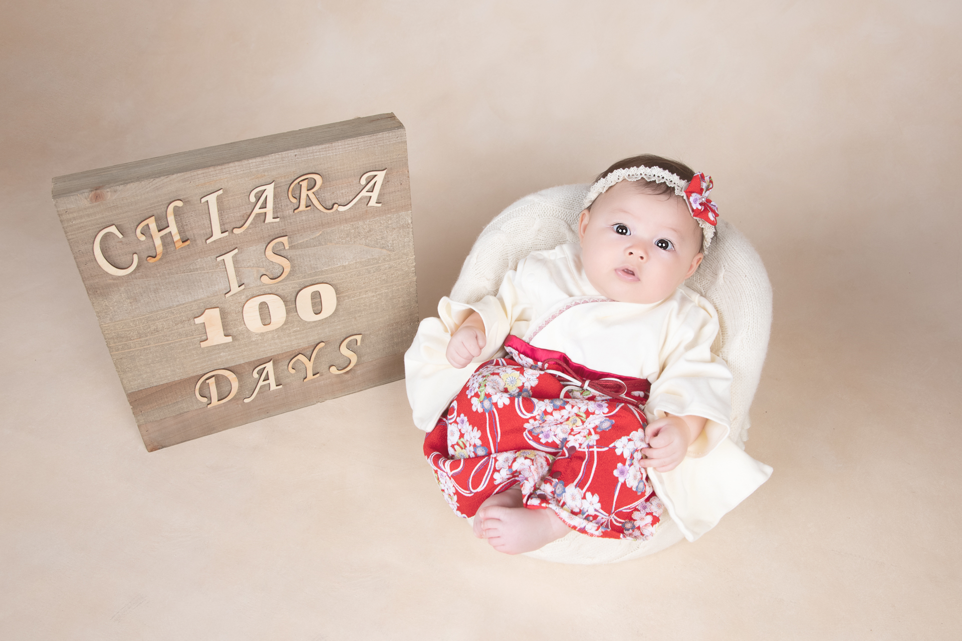 Baby girl rest on light colored prop wearing light colored and red dress and headband. Sign next to her shows "Chiara is 100 days"
