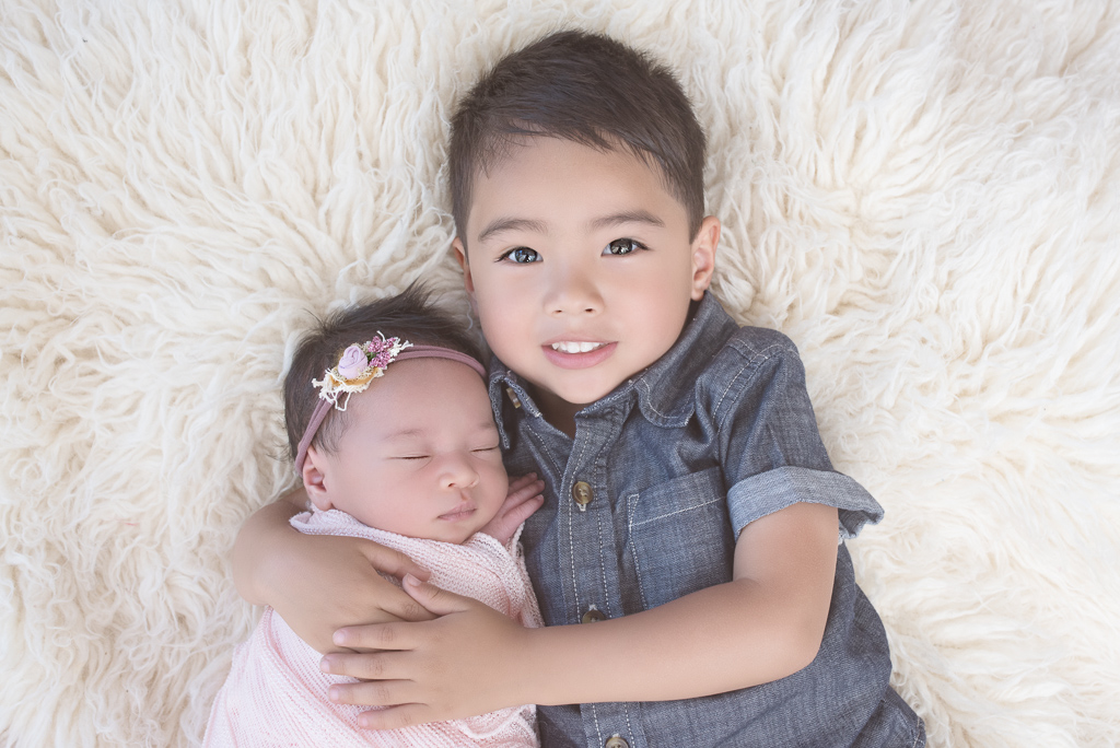 Big brother wearing blue T-shirt outfit hugs newborn sister wearing pink headband and wrap. Fluffy light color carpet decorates the background.