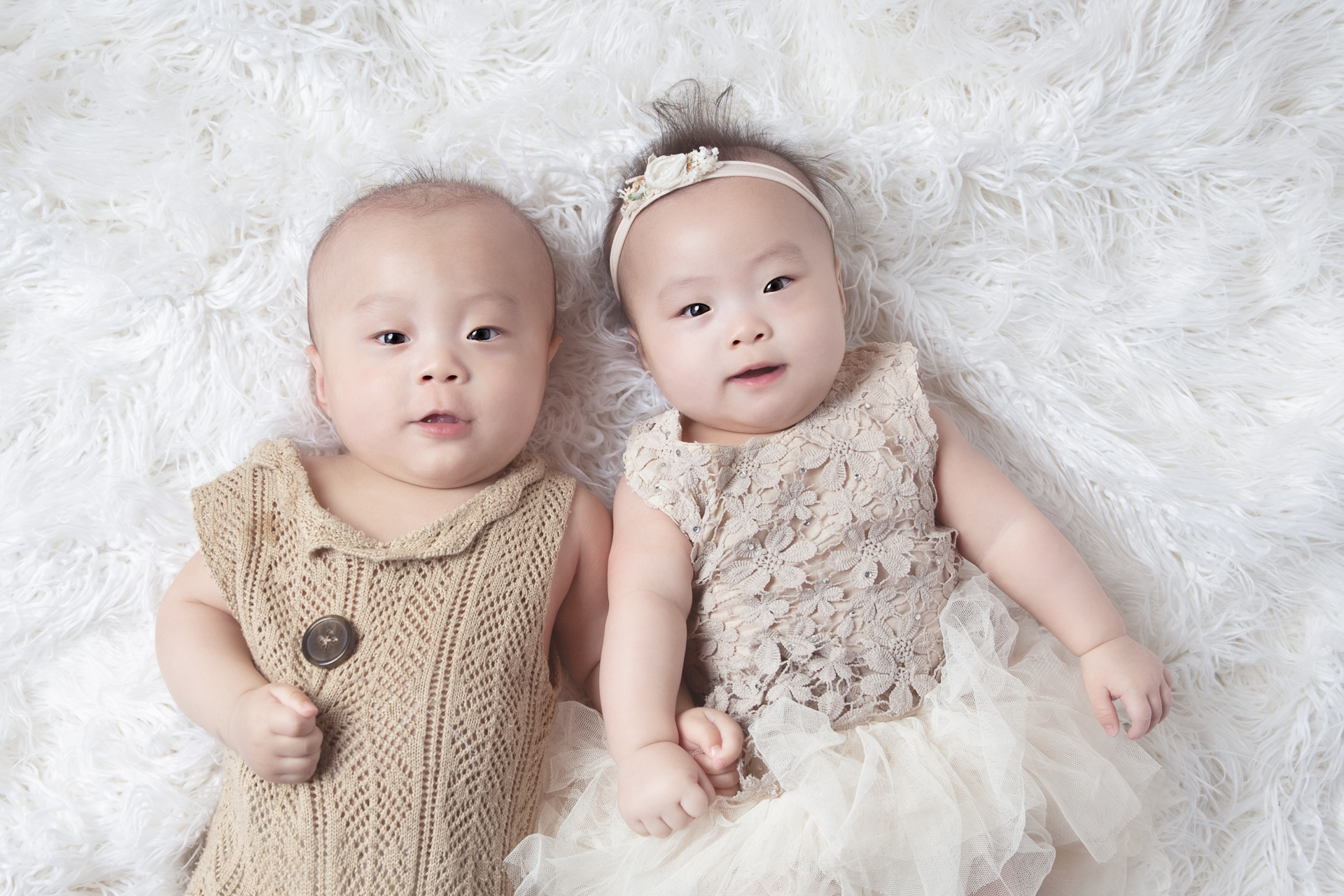 6 month old sister and brother resting on white fluffy backdrop on light brown tones outfits look at the camera.