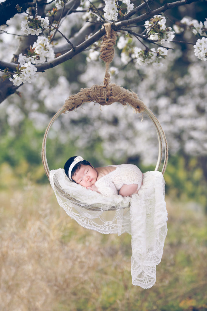 Newborn rests outdoors on hanging prop while wearing white outfit and white headband. White flowers decorates the scene.