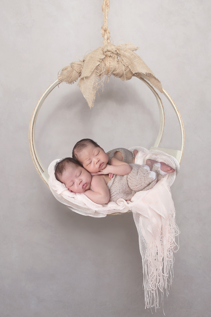2 newborn siblings rest together on a hanging prop wearing light brown outfits