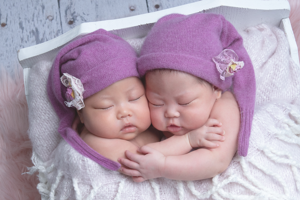 2 newborn siblings rest together while wearing purple hats on a bed prop, hugging each other