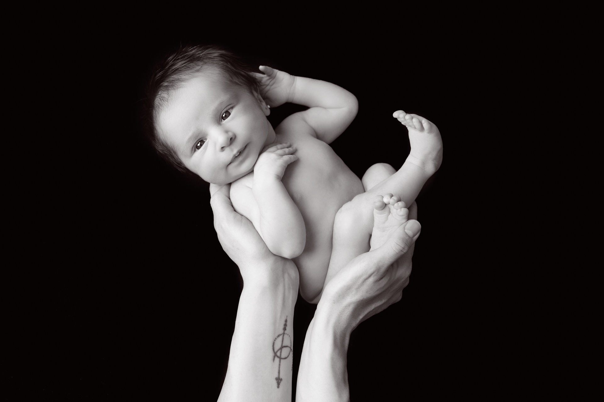 Newborn girl is held by parents by one of each parents hands. Tattoo on wrist. Black and white image.