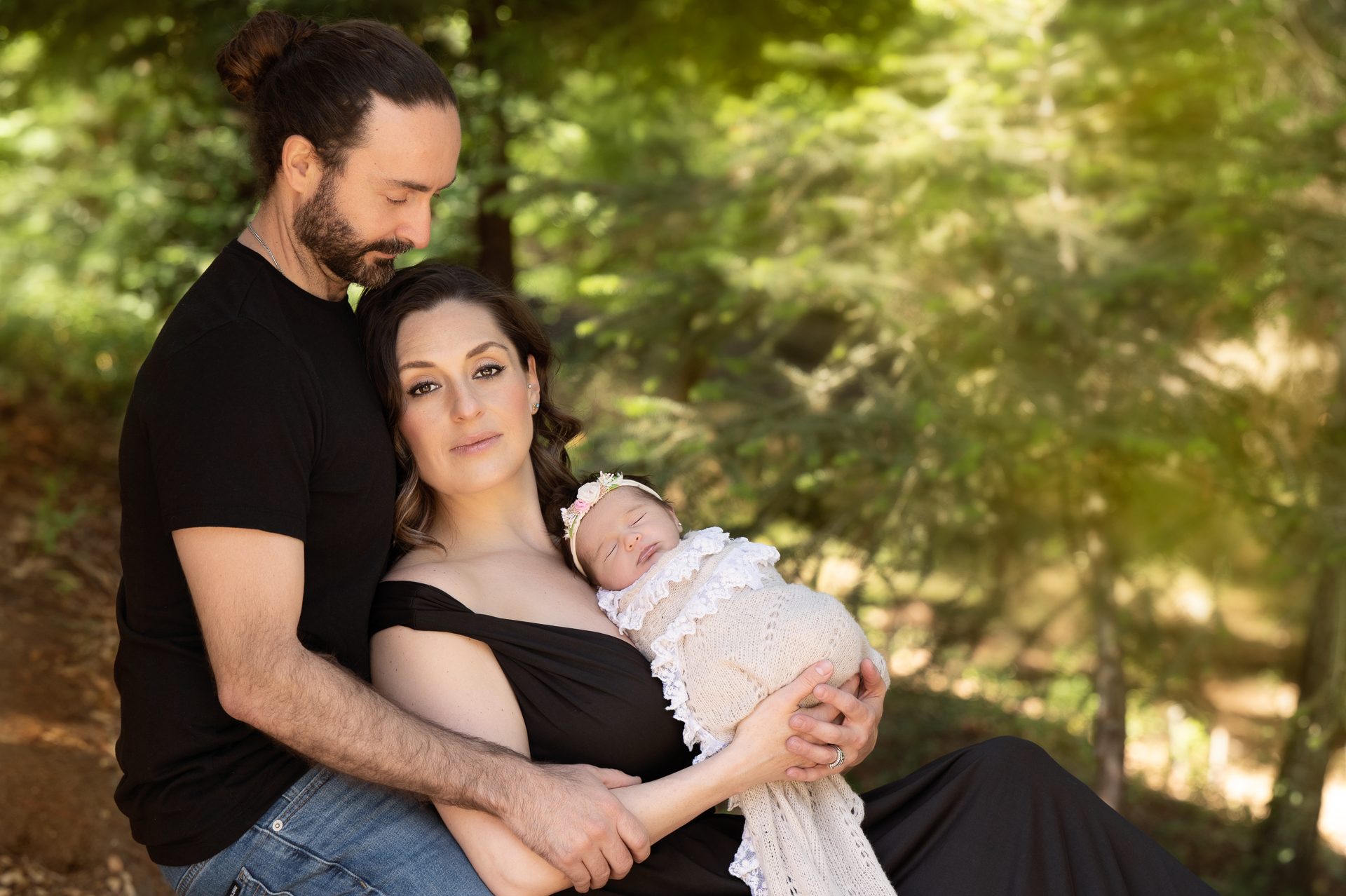 Couple wearing black outfits posing outdoors with their newborn daughter. Baby wear light color wrap and headband.