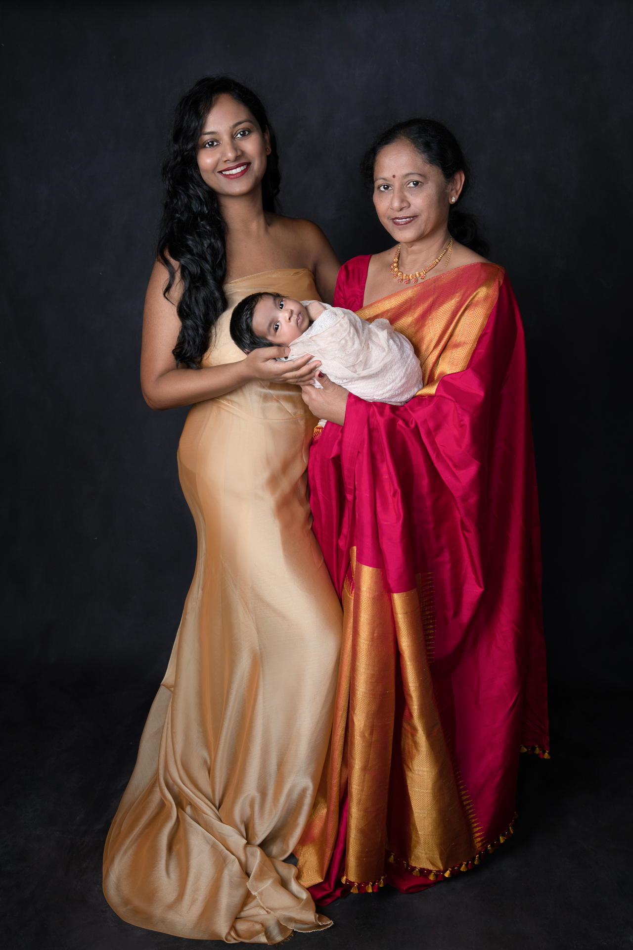 Grandmother, daughter and newborn girl posing indoor. Black backdrop. Grandmother wears sari. Mom on golden color dress. Baby on light color wrap.