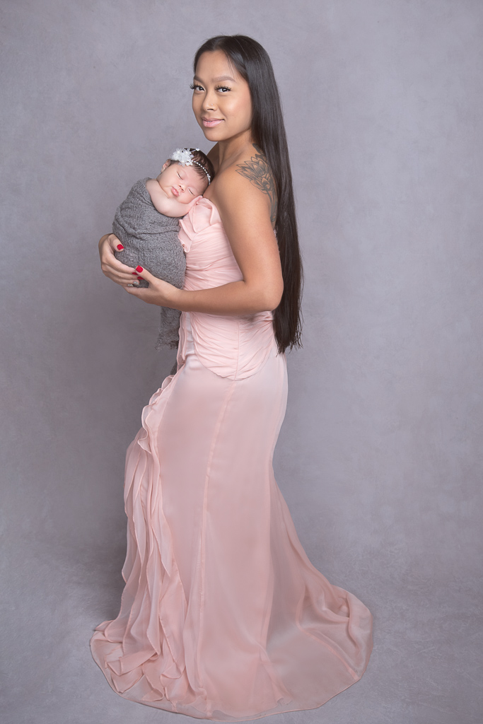 Woman on pink dress posing indoors with her newborn daughter, which wears gray wrap outfit and a white headband. Light gray backdrop.