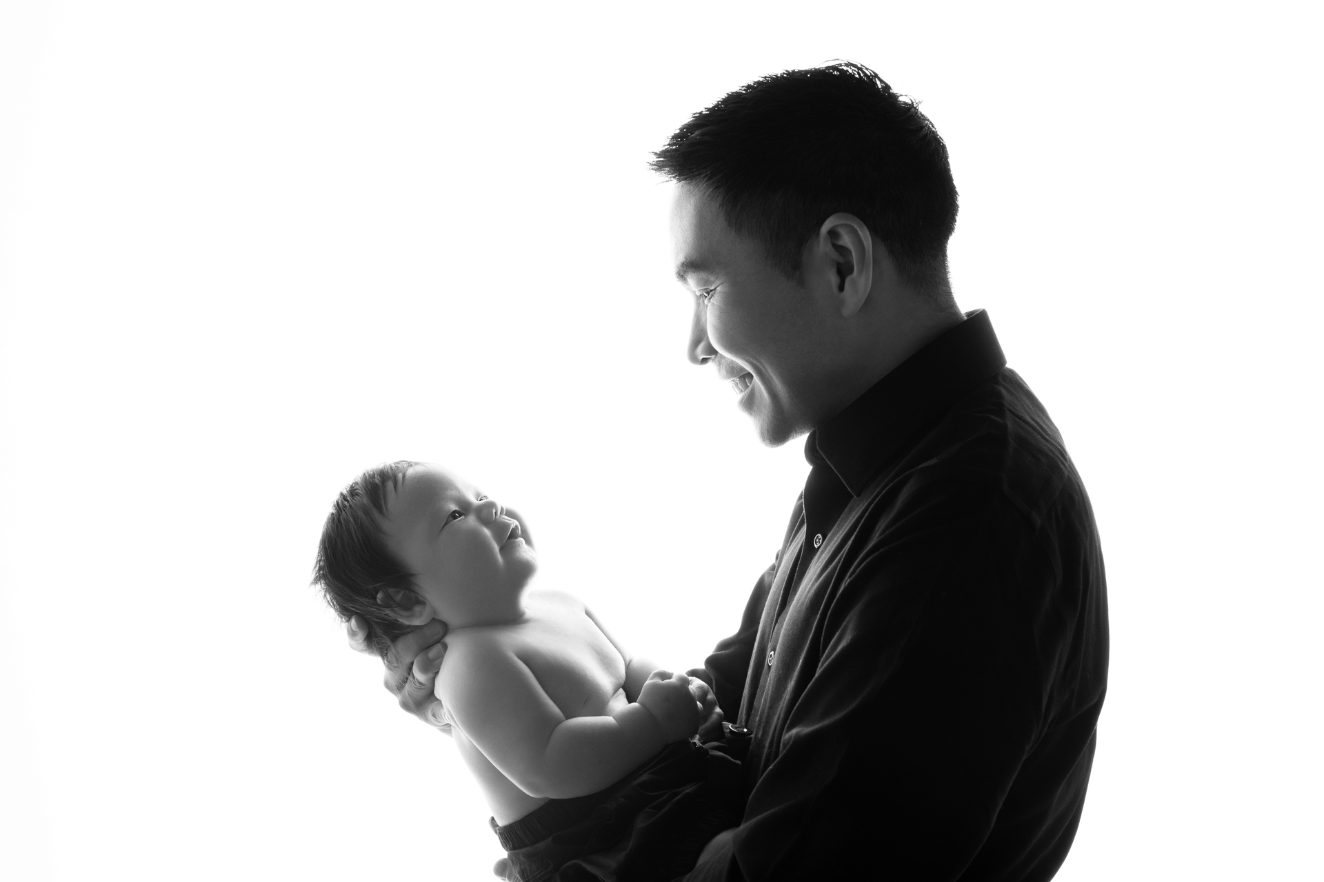 A father poses indoors with his son, white background. Dark outfits, black and white image.