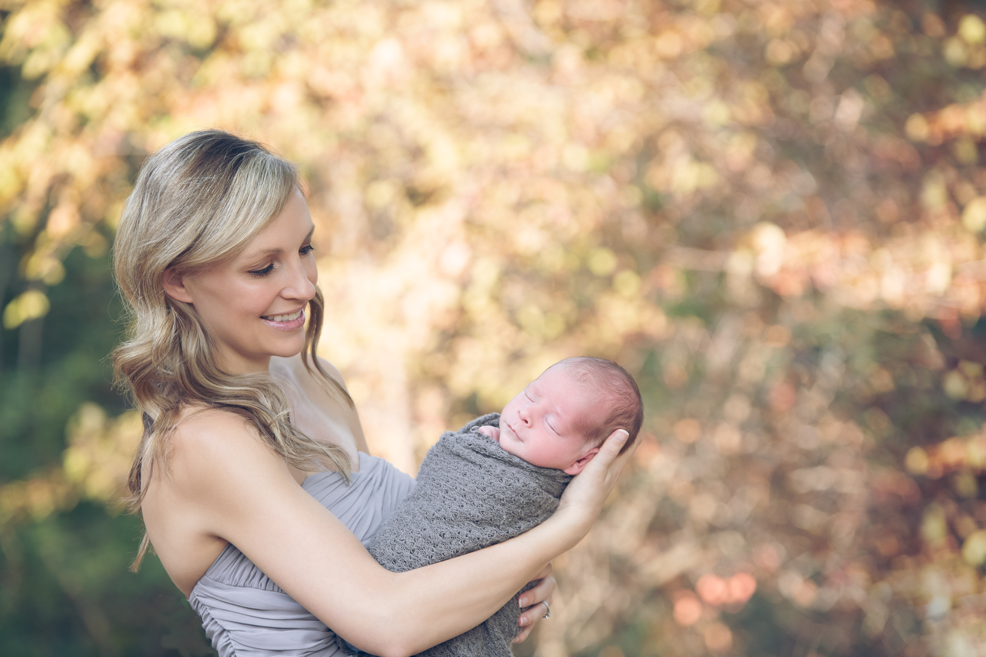 Woman on a gray dress holds her newborn son during fall season outdoors.