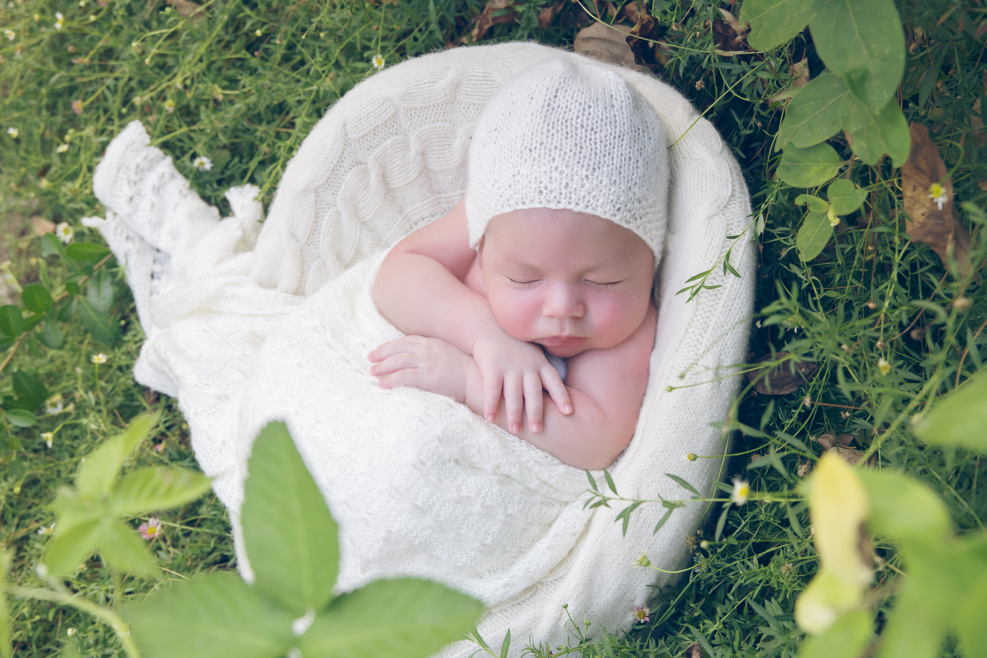 Newborn wearing light beige hat and light beige color wrap rests on light beige round prop outdoors.