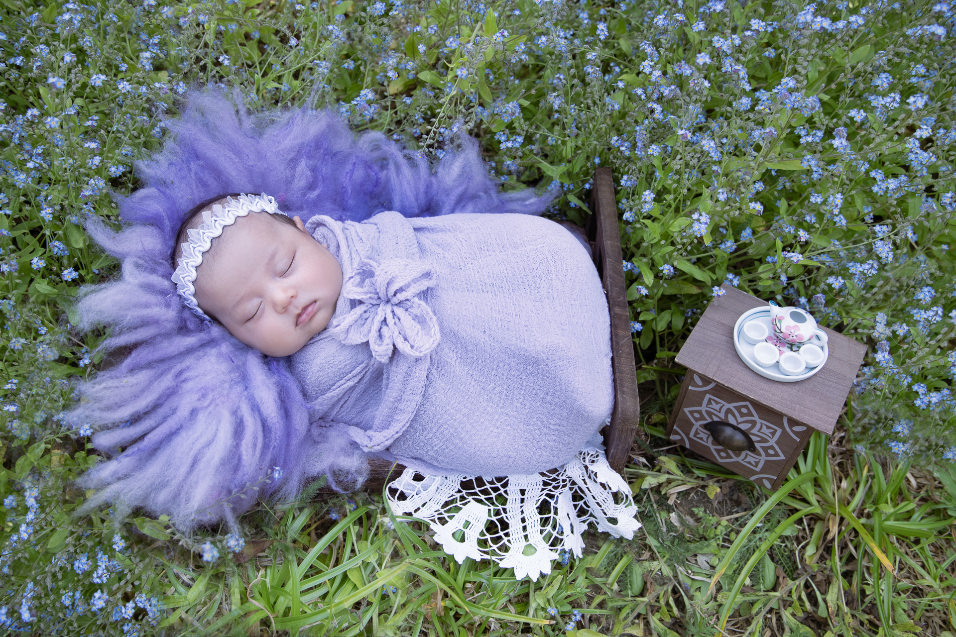 Newborn girl rest on wood bed prop outdoors. Purple wrap, white headband. Purple flowers sourrounding her. Teapot and tea cups on small table decorating the scene.