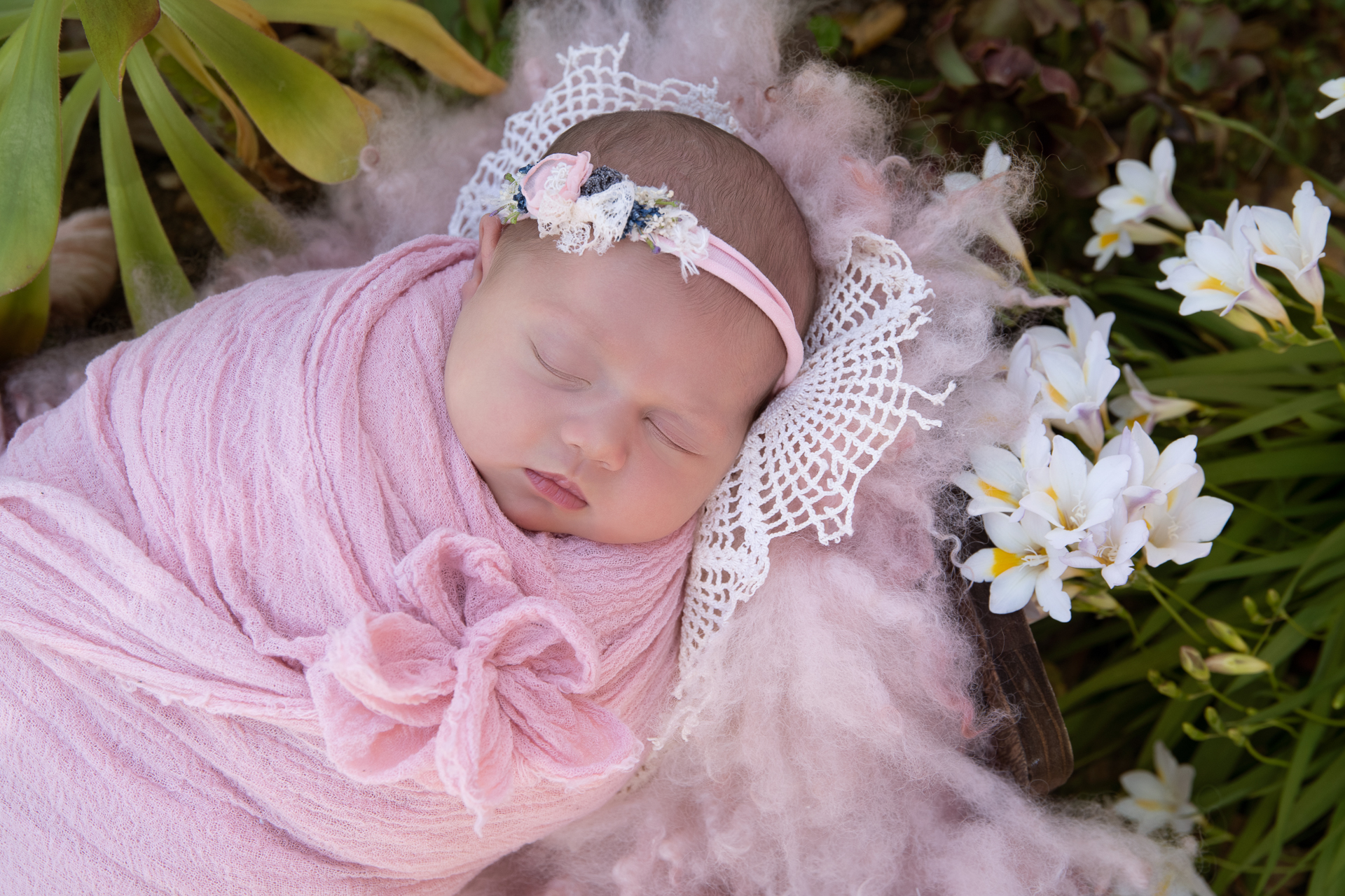 Newborn baby girl wears pink wrap and headband while resting on prop outdoors