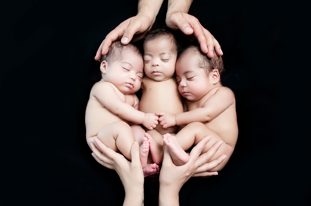 3 newborn siblings rest together in the hands of their parents. Black background.