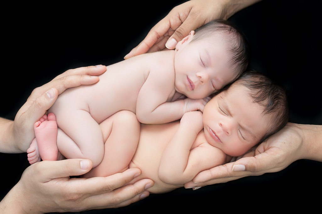 two newborn siblings rest together on their parent's hands. black background