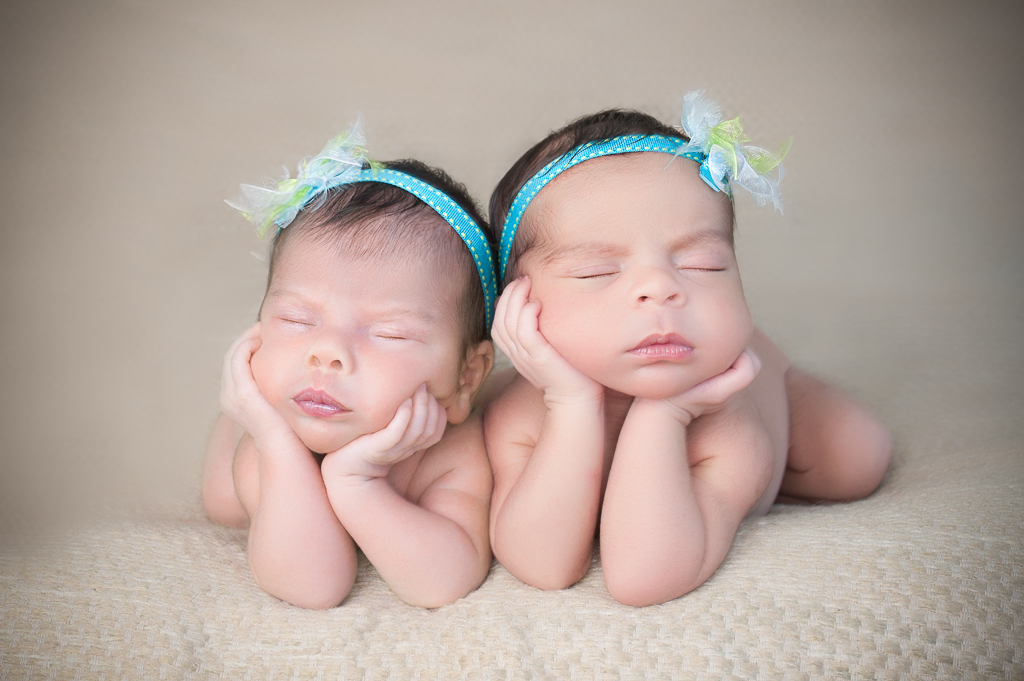two newborn babies rest on their own hands on froggy pose. light blue headbands. light backdrop