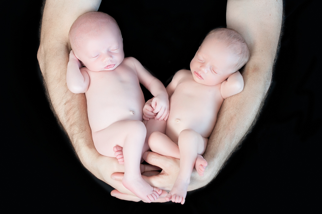 Two newborn babies rest on their father arms and hands. Black background