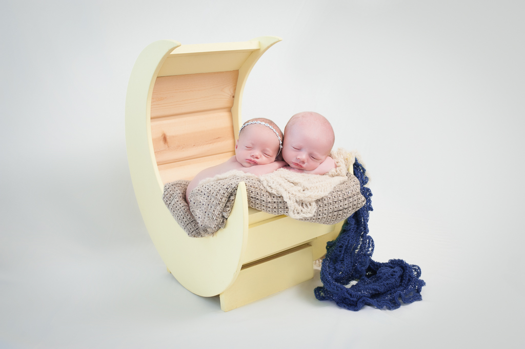 Two newborn babies rest together on a moon shaped prop, light background, of of them wearing a headband