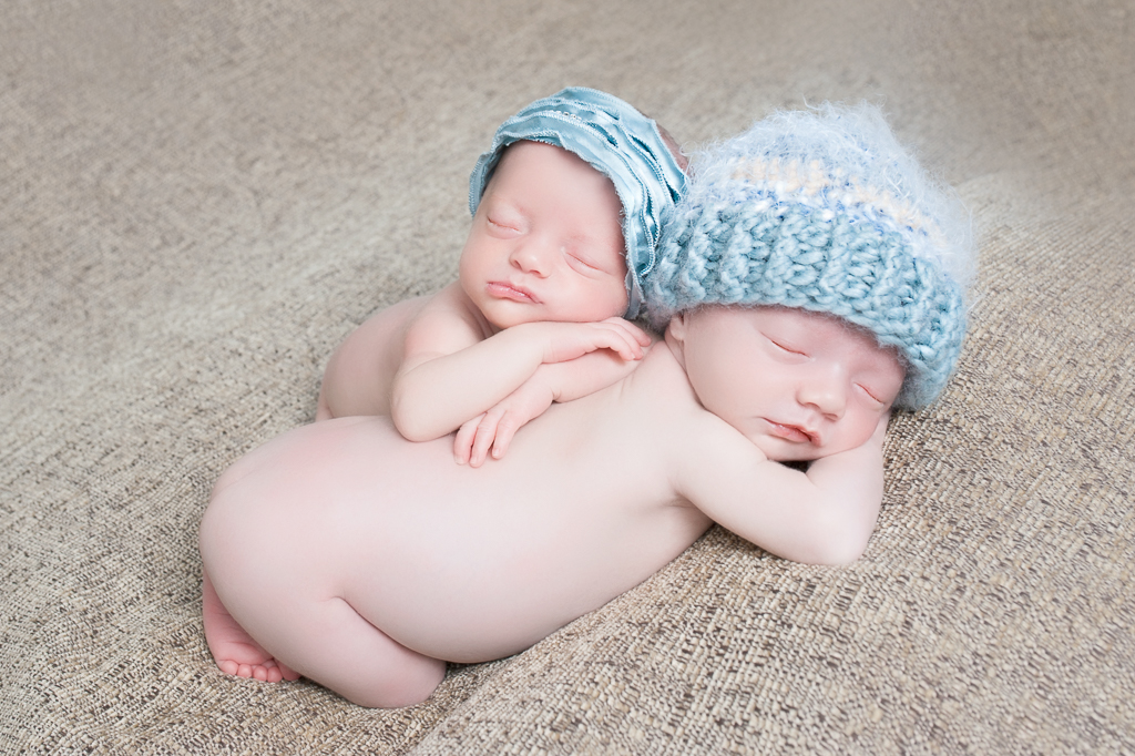 two newborn siblings wearing a blue hat and a blue headband rest together, one rests on top of the other