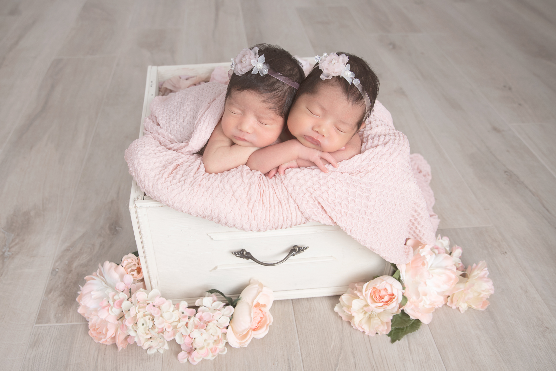 Two newborn sisters posing on a square basket prop wearing pink headbands, flowers decorate the scene