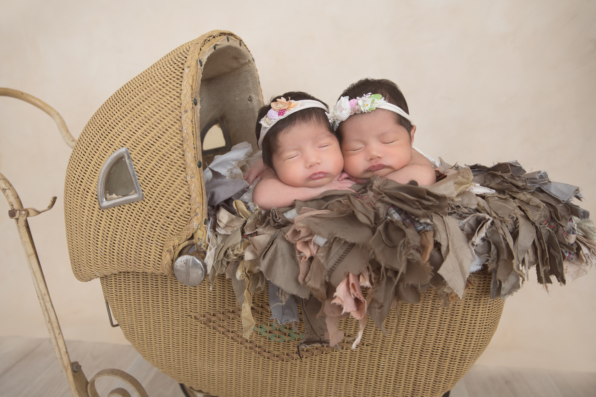 2 newborn sisters rest together in a old fashion decorative brown colored stroller, light brown backdrop, light colored headbands