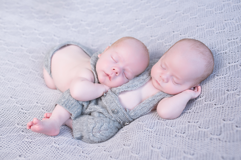 2 newborn siblings sleeping together on gray outfits, one of them is resting his head on his sibling torso. Gray background, straight on angle shot