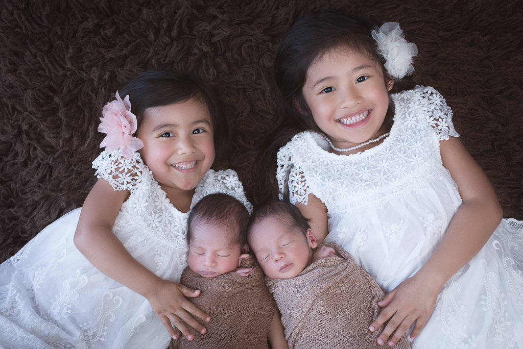 2 girls smile at the camera while holding their newborn brothers. both wearing white dresses, babies wrapped on brown tones. brown fluffy carpet background