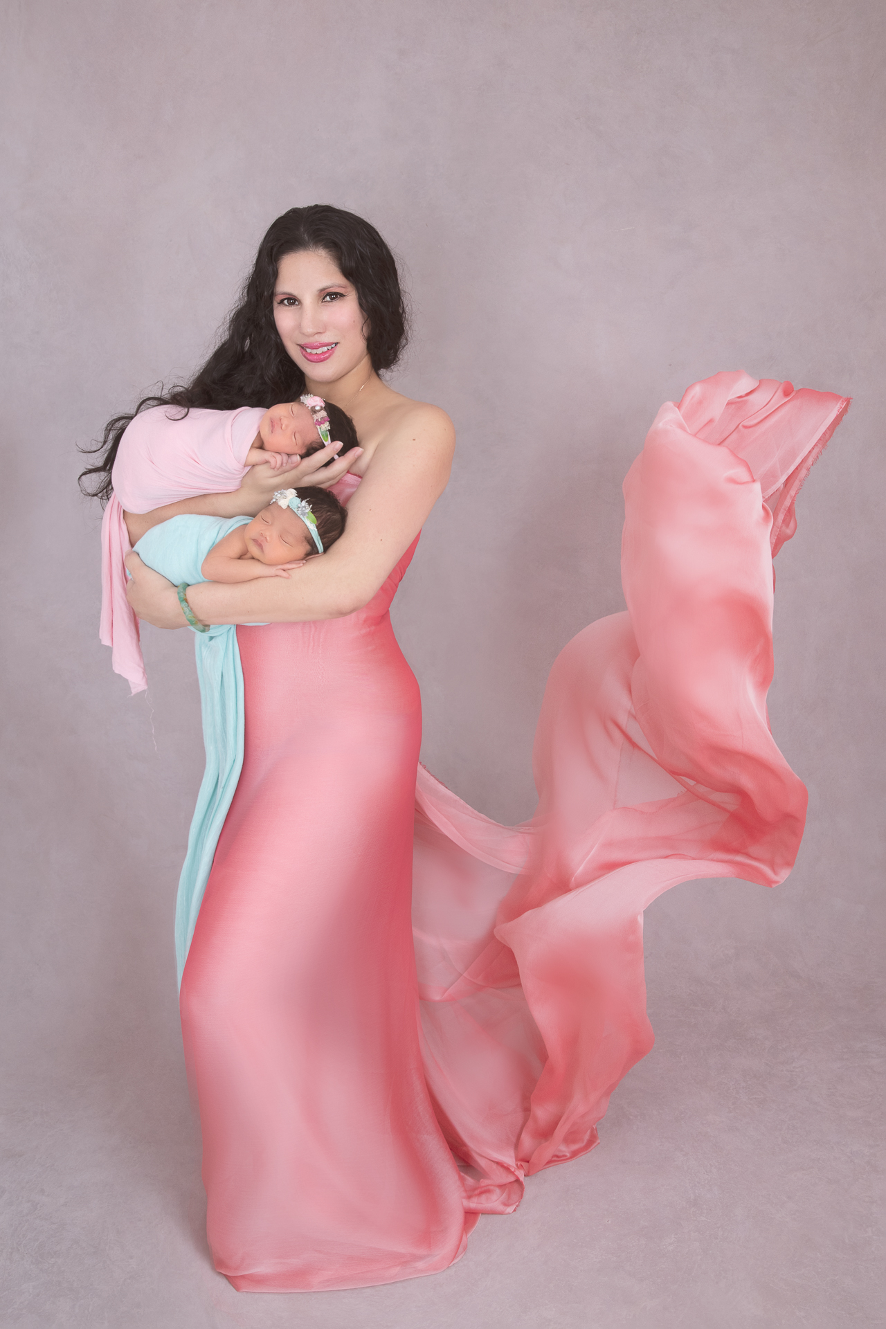 Mom posing indoors with her 2 newborn daughters on pink and green tones.