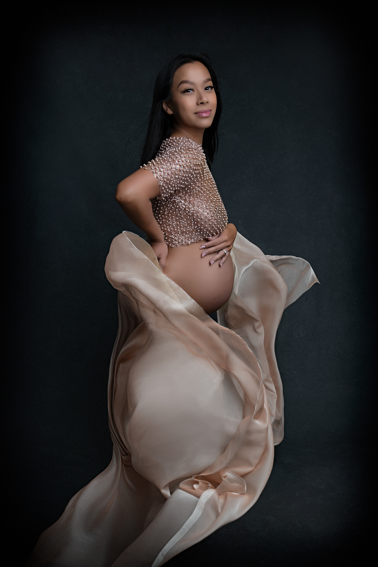Pregnant woman wears pearl top and light brown fabric on dark backdrop