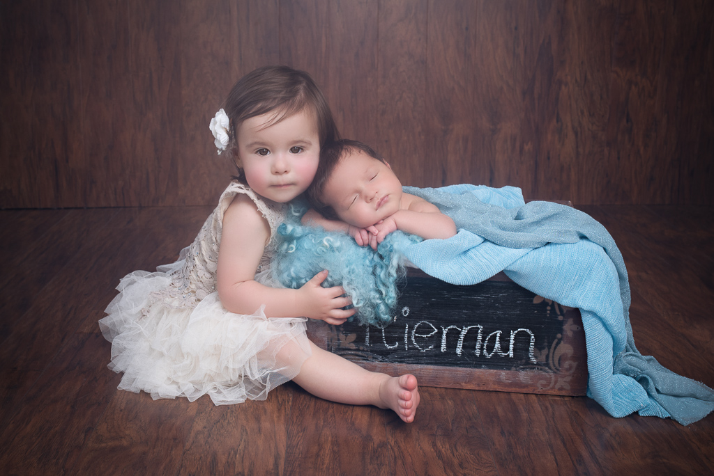 Toddler sister sits next to her newborn brother which rests on a square prop basket. Brown background