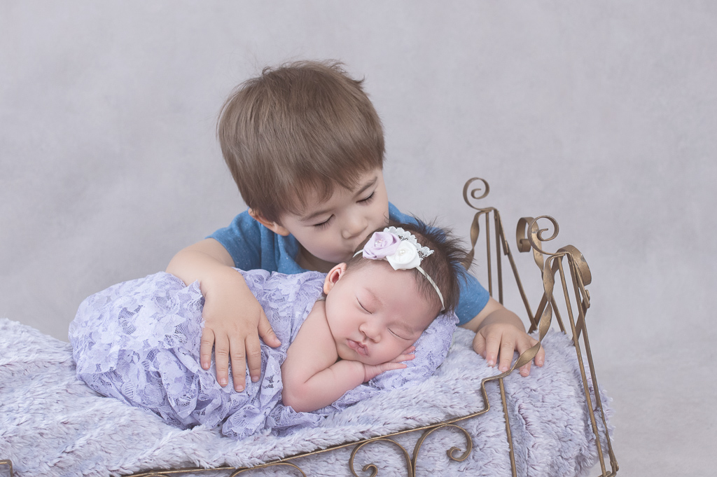 Toddler on blue T-shirt kisses his newborn sister on the head while she rests on a prop bed. Baby wears white and pink headband and purple wrap.