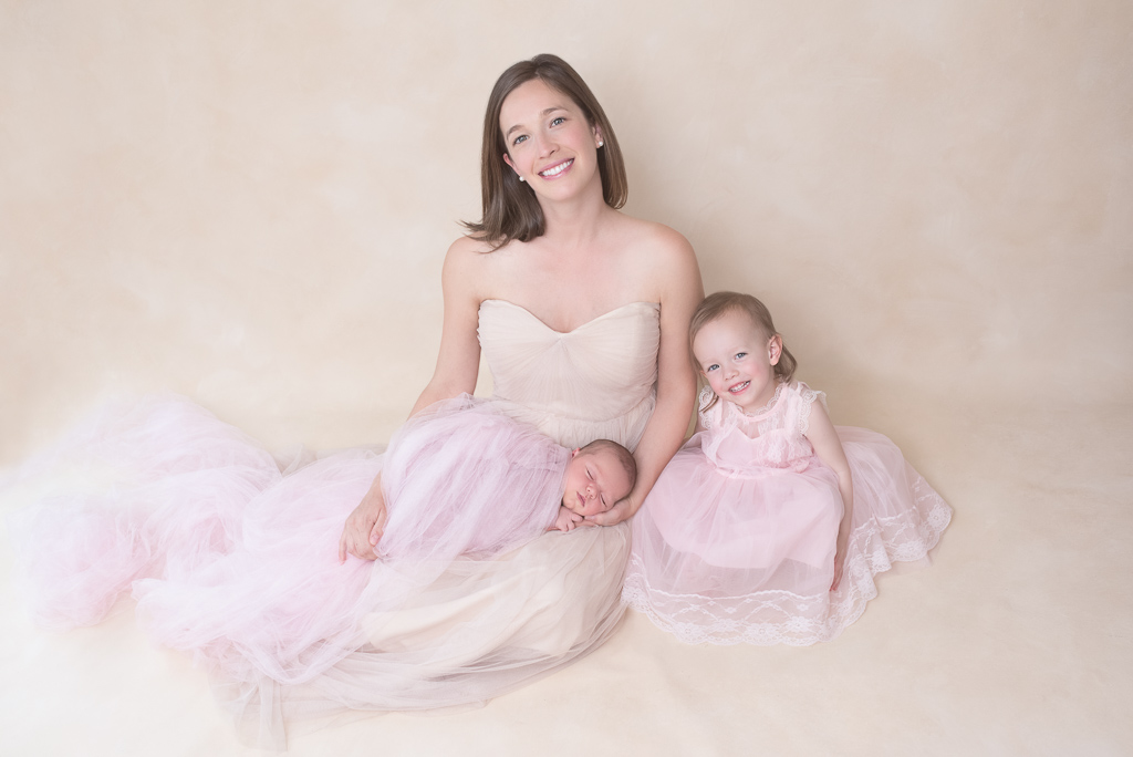 Mother sits next to her toddler daughter while holding her newborn baby girl. Kids on pink colors, mom on light brown dress. Light brown backdrop.