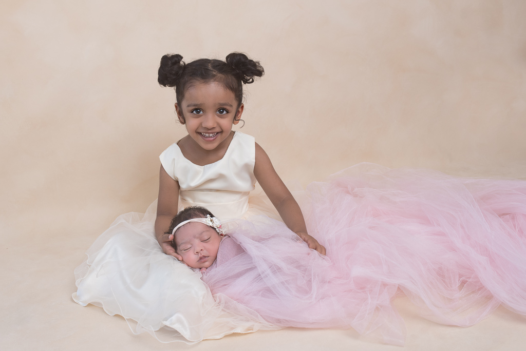 Older sister in light color dress sits while holding her newborn sister that wears light colored headband and pink fabric. Light brown backdrop.