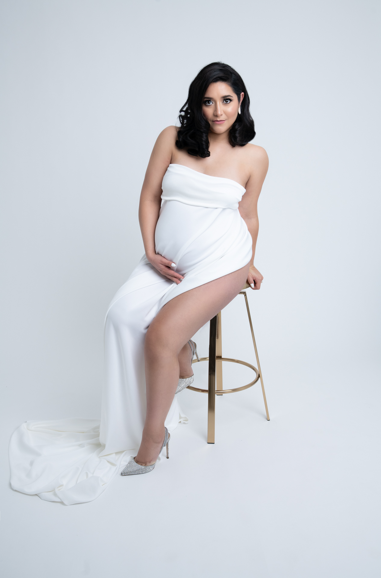 Dark hair pregnant woman sitting down on golden color chair wearing white fabric dress