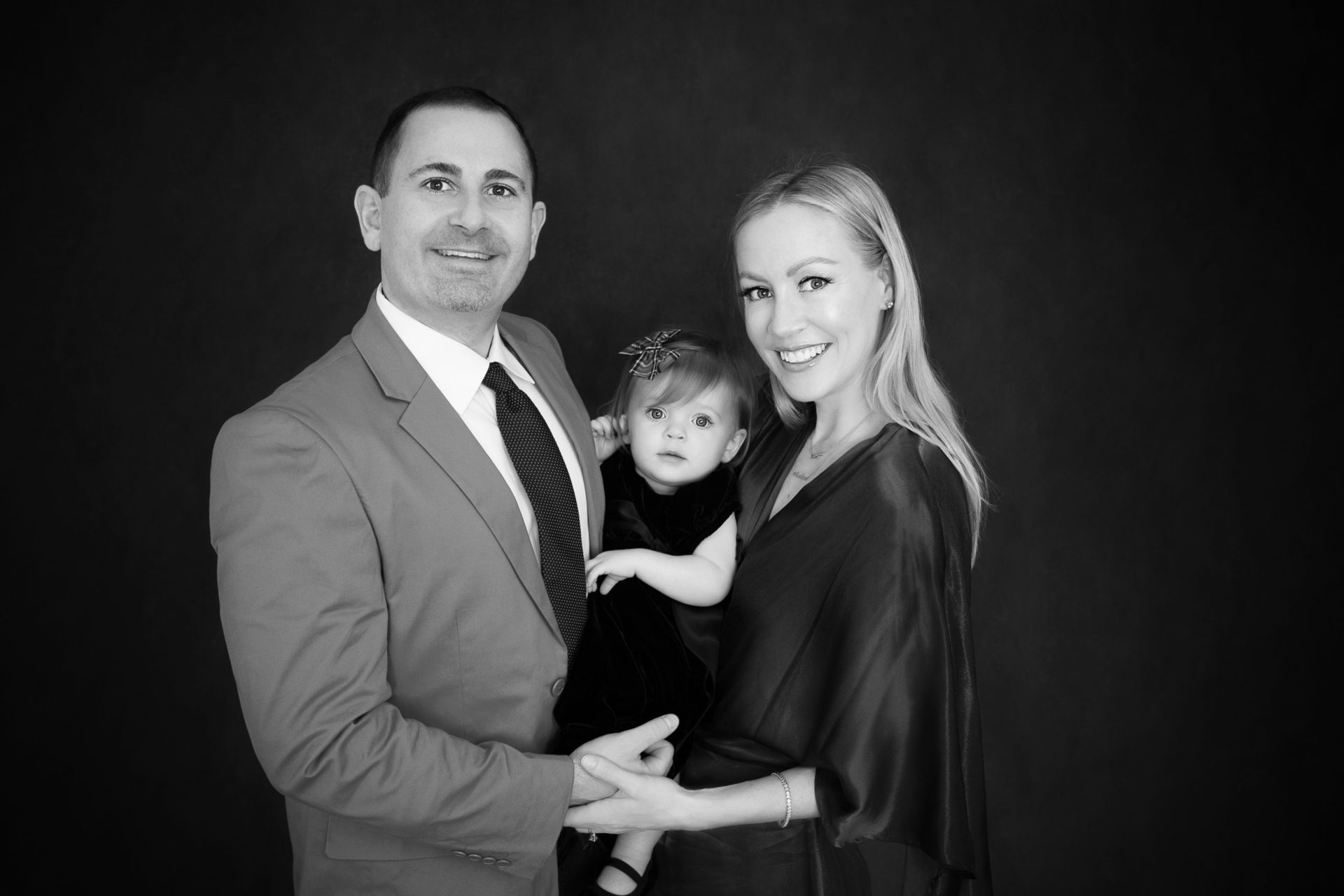 Family black and white photo shows father, mother and one year old daughter posing indoors. Father wear a tie, mom and daughter in dresses.