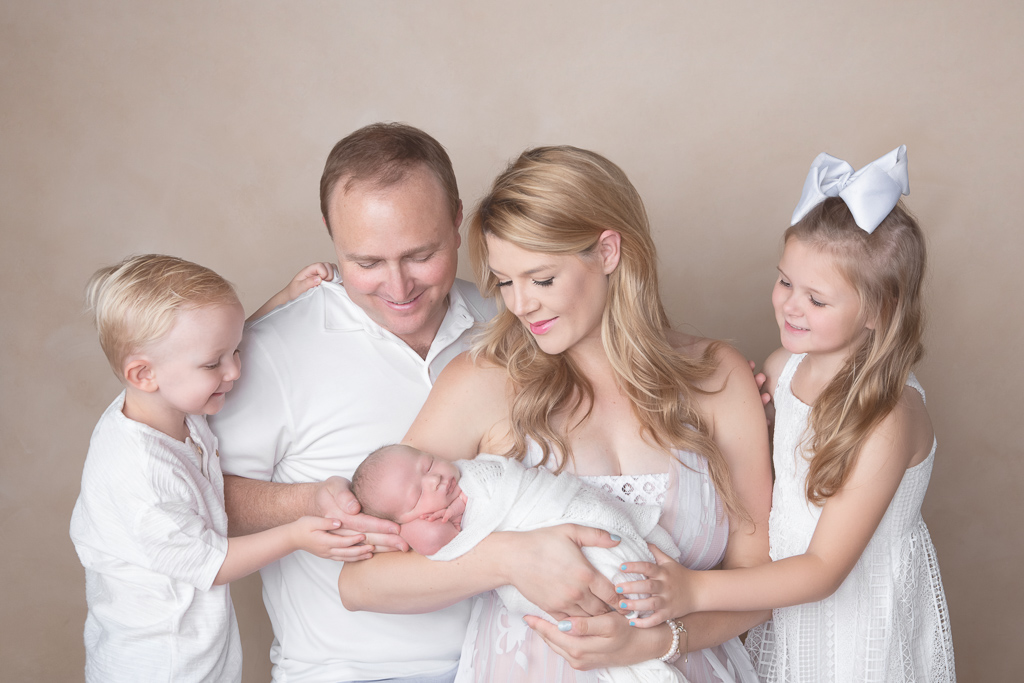 Family of 5 poses indoor on a light brown background. Dad, mom, 2 year old boy, 6 year old girl and newborn boy.