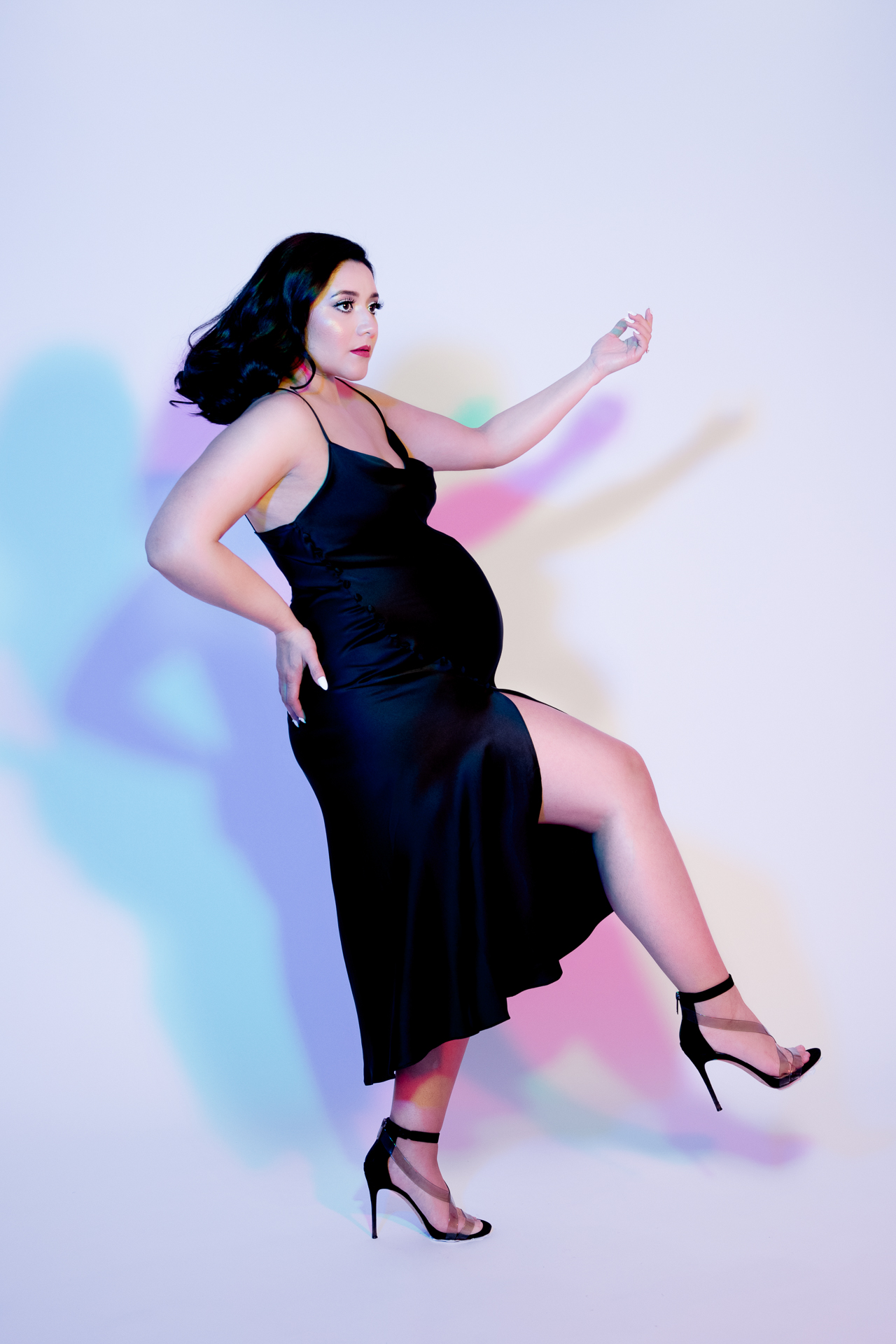 Pregnant woman wearing black dress and black high heels. White backdrop reflects colorful shadows.
