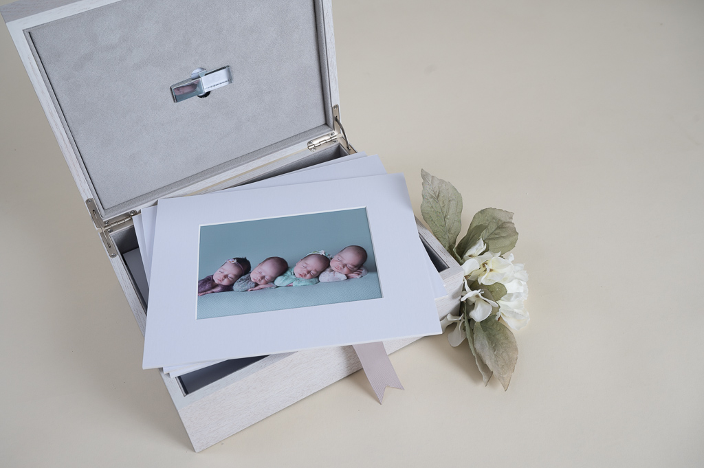 Open white photo box shows 4 newborn siblings frame and a pen drive containing negatives digital information. Flowers decorating.