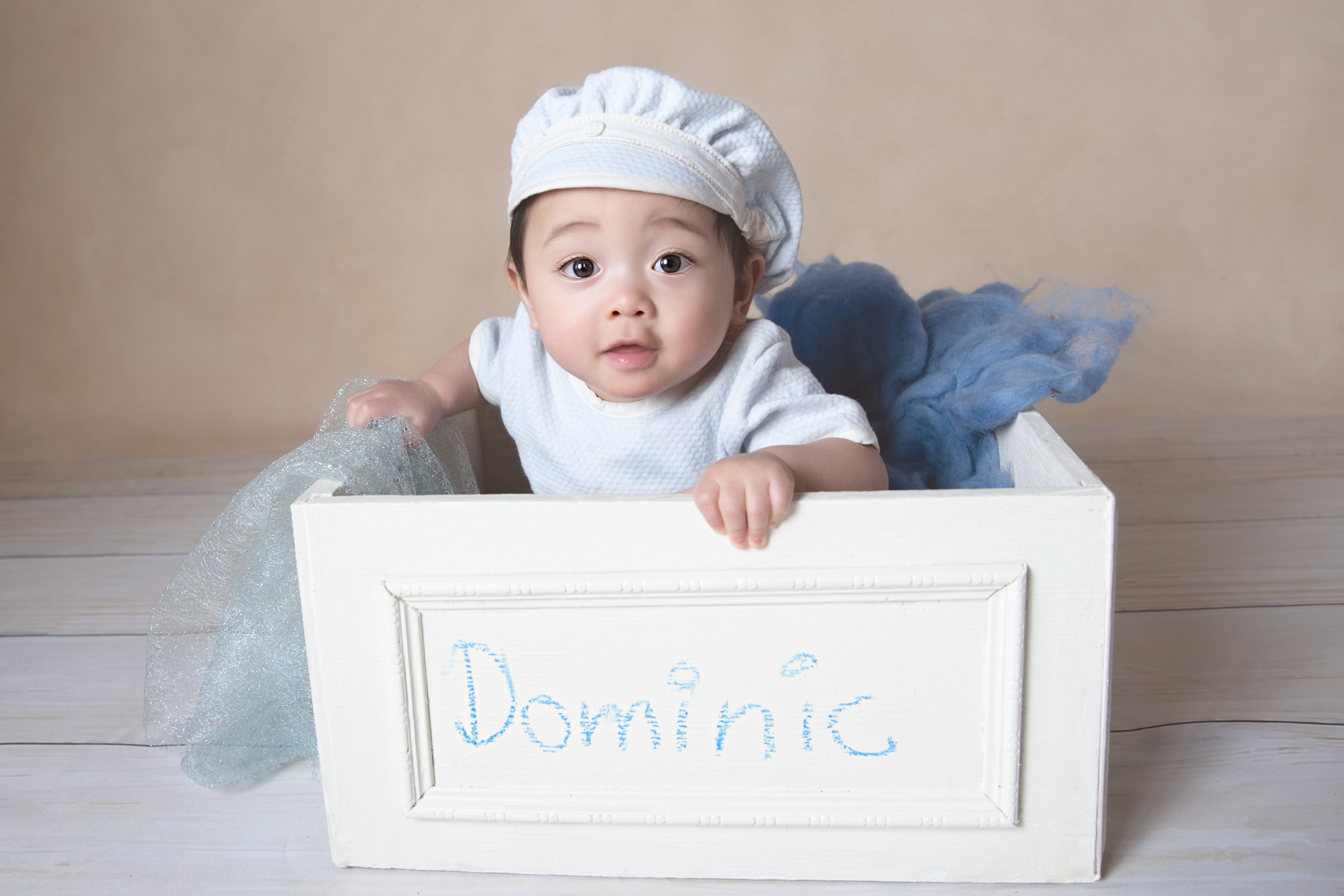 Baby boy sitting on prop wears a blue hat , light brown backdrop