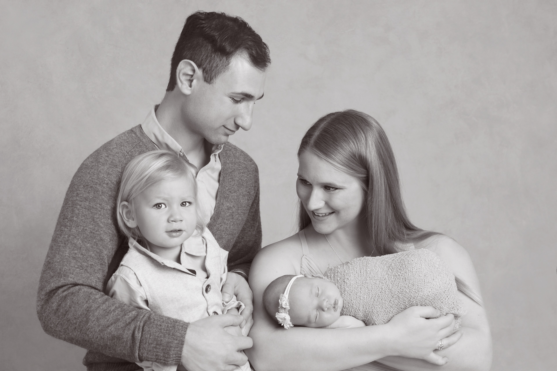 Family of 4 posing indoors in a black and white image. Dad, mom, toddler, newborn girl.