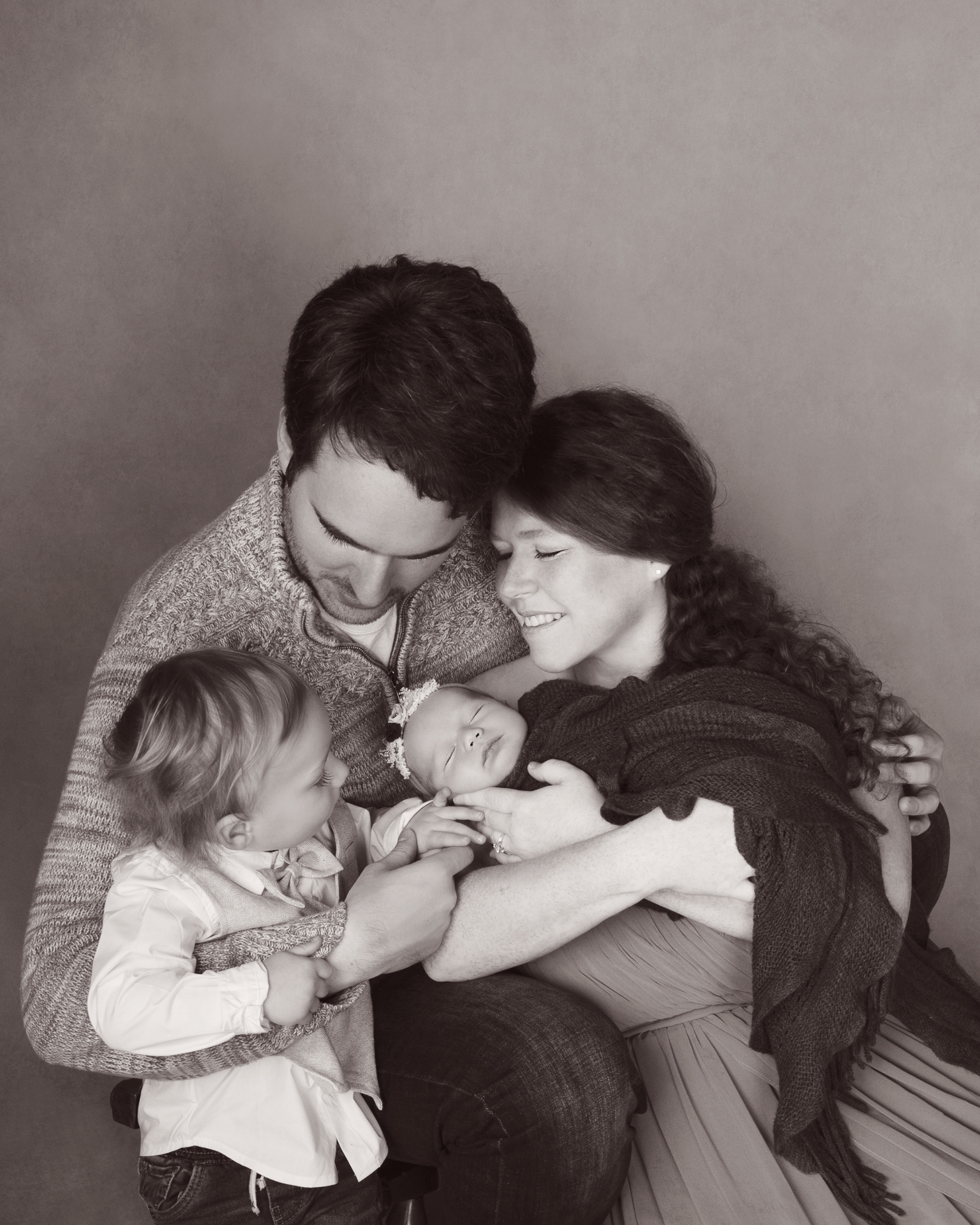 Family of 4 posing indoors. Black and white image. Dad, toddler boy, mom and newborn baby girl.