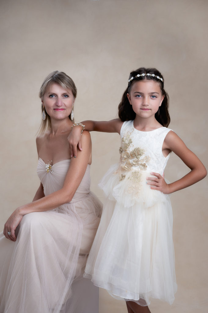 Mother and 9 year old daughter posing indoors on cream color and white dresses, respectively.