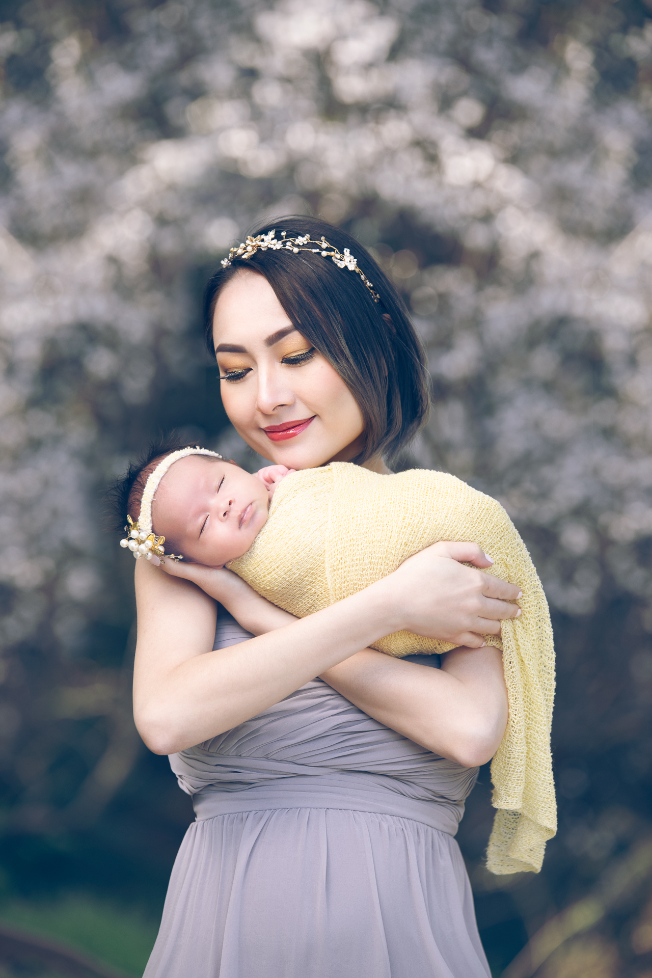A mother on a gray dress poses outdoors with her newborn daughter which wears yellow wrap outfit and yellow headband.