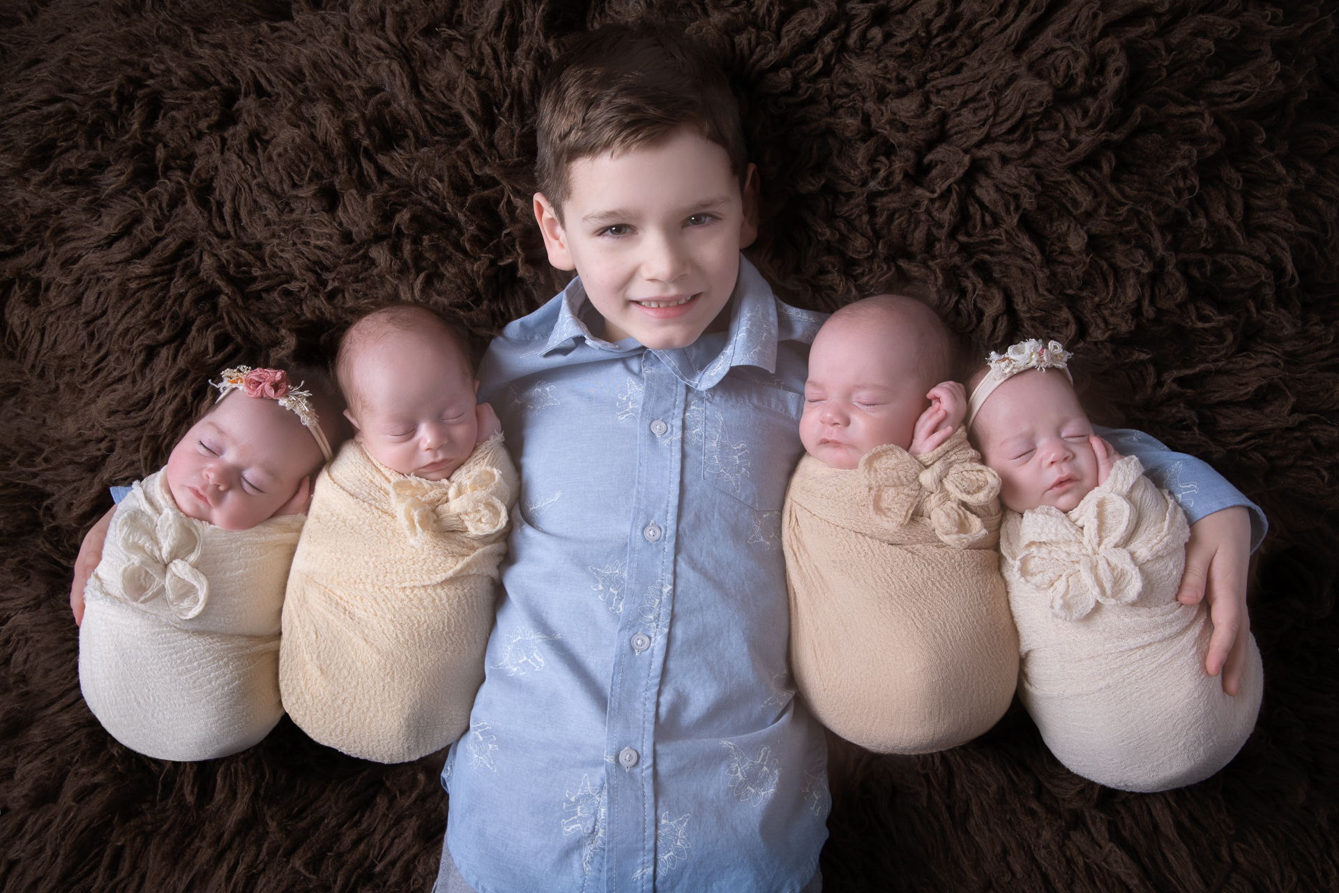 Big brother holds his 4 newborn siblings. Two babies on each side. Dark brown carpet as background.