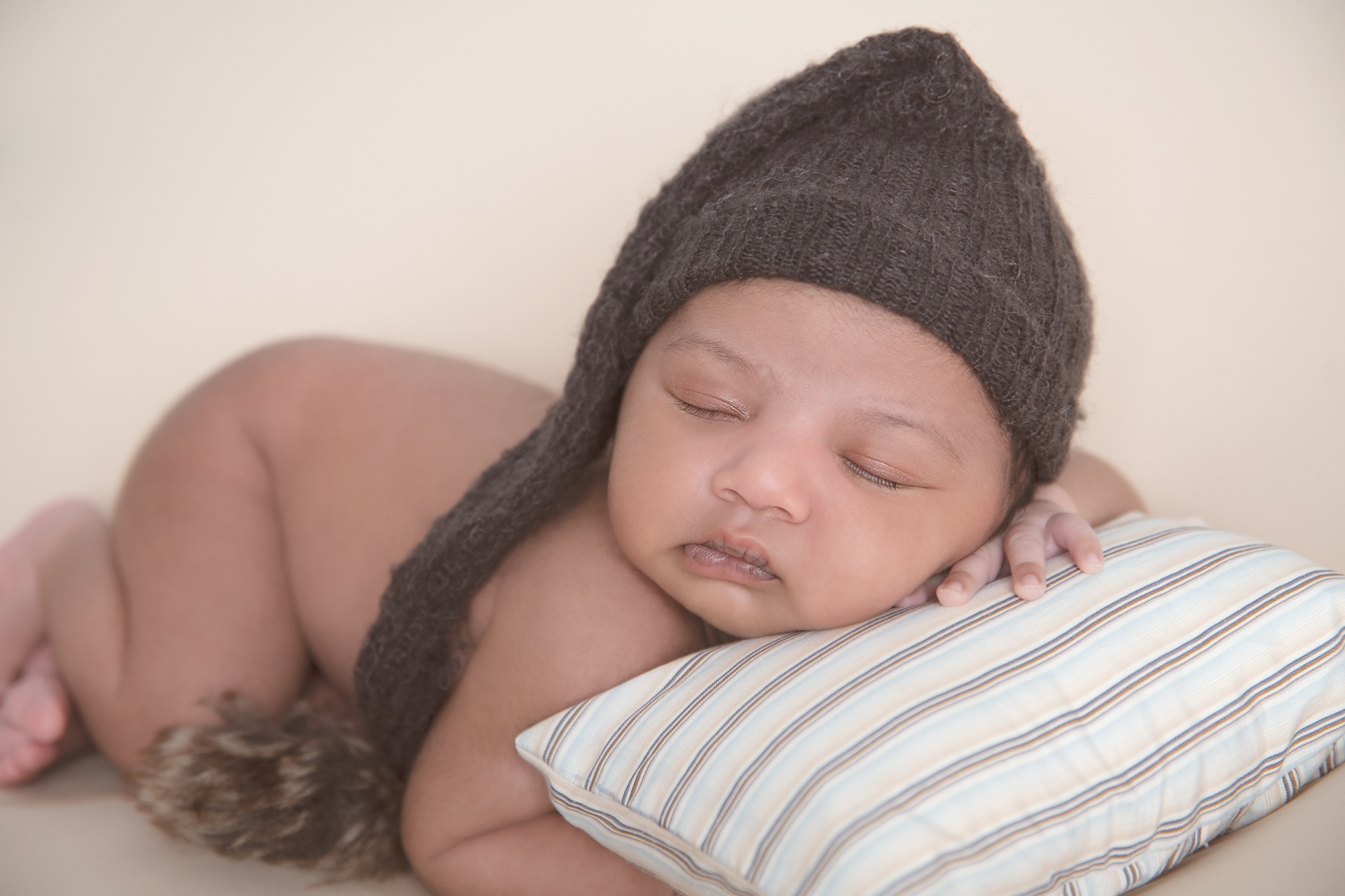 Newborn baby wearing brown hat rests on a small pillow.