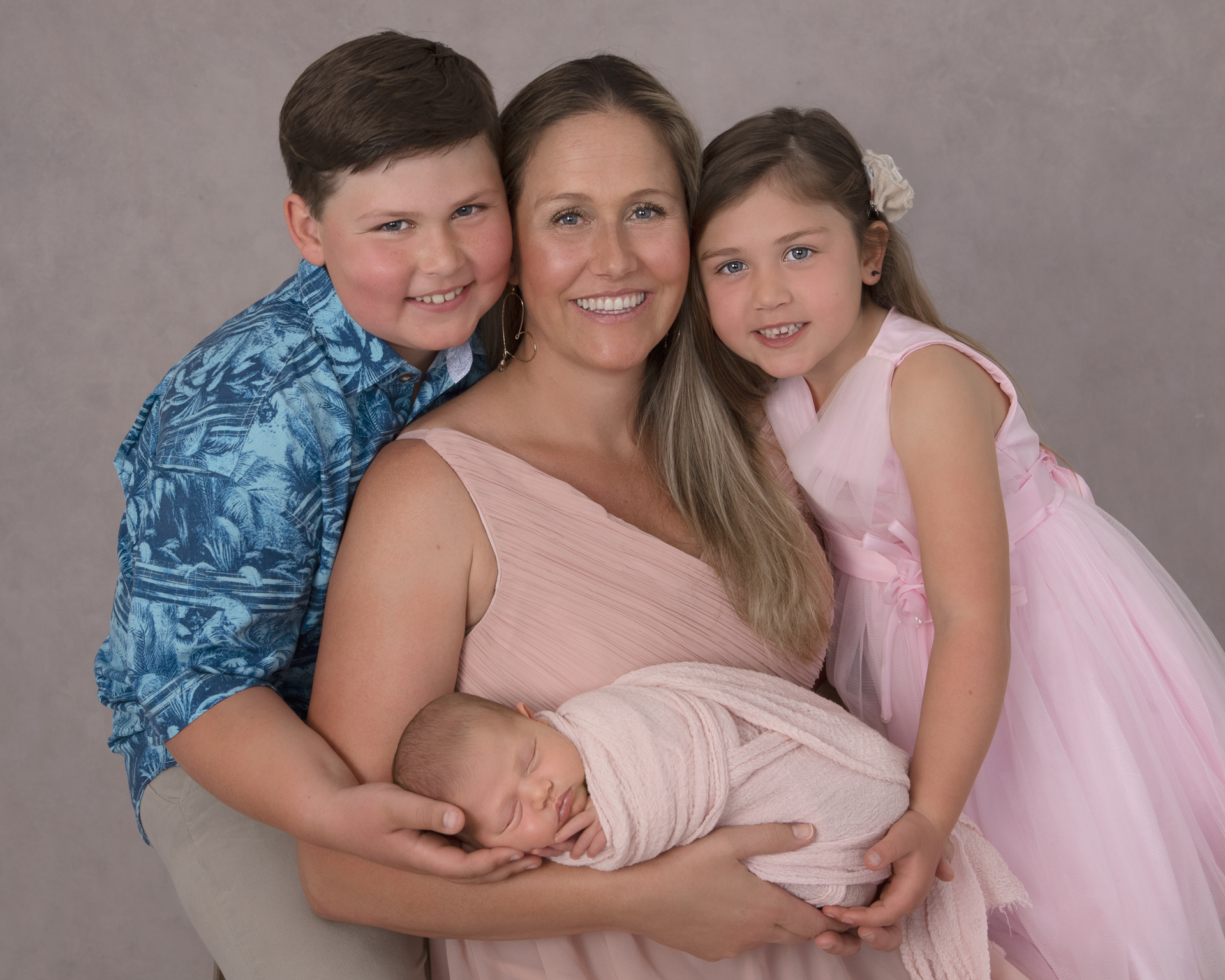 Mother posing indoors holding her newborn daughter, accompanied by her 2 older kids. Girls on pink, boy on blue outfit. Gray backdrop.